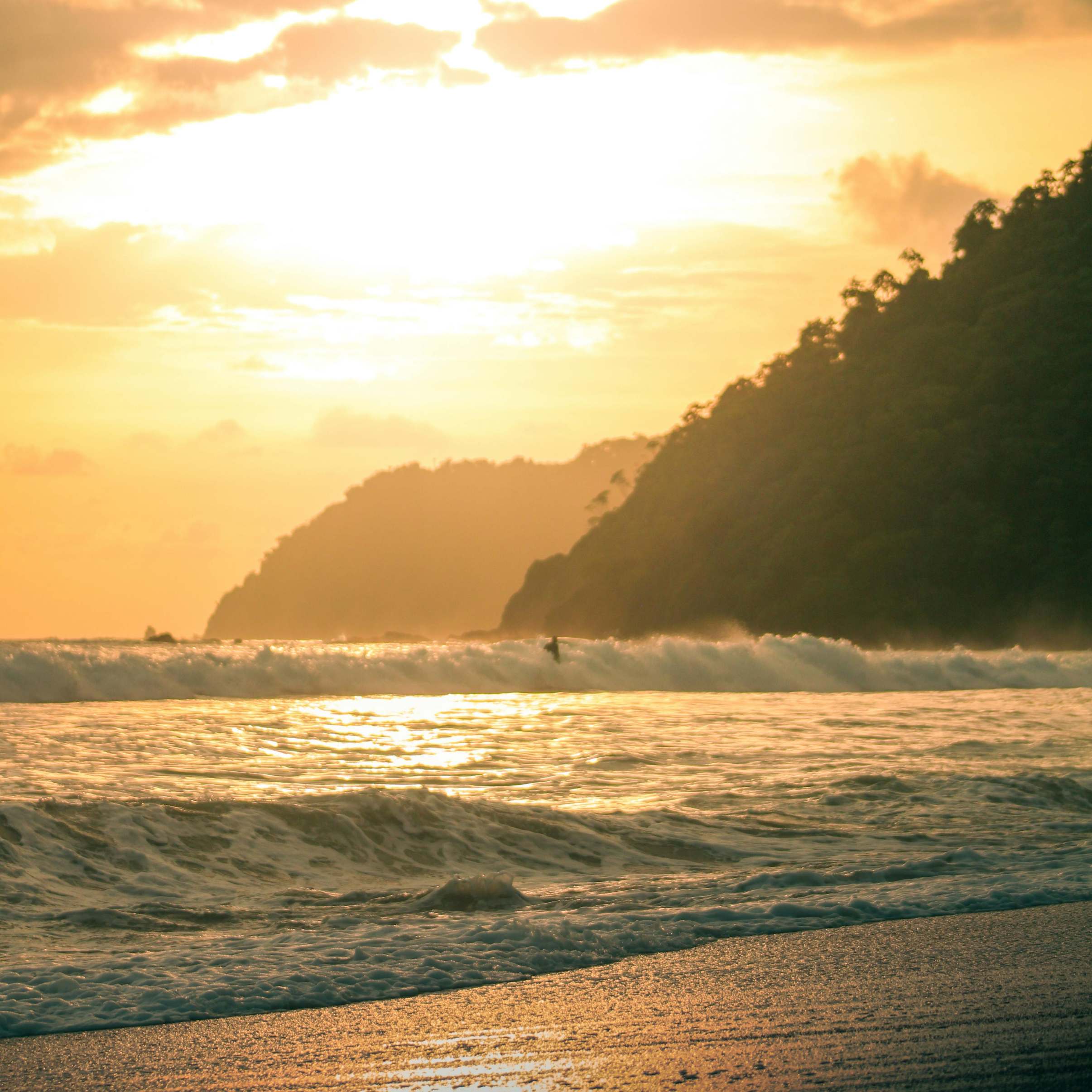 Ocean waves at sunset with a surfer and tree-covered hills in the background.