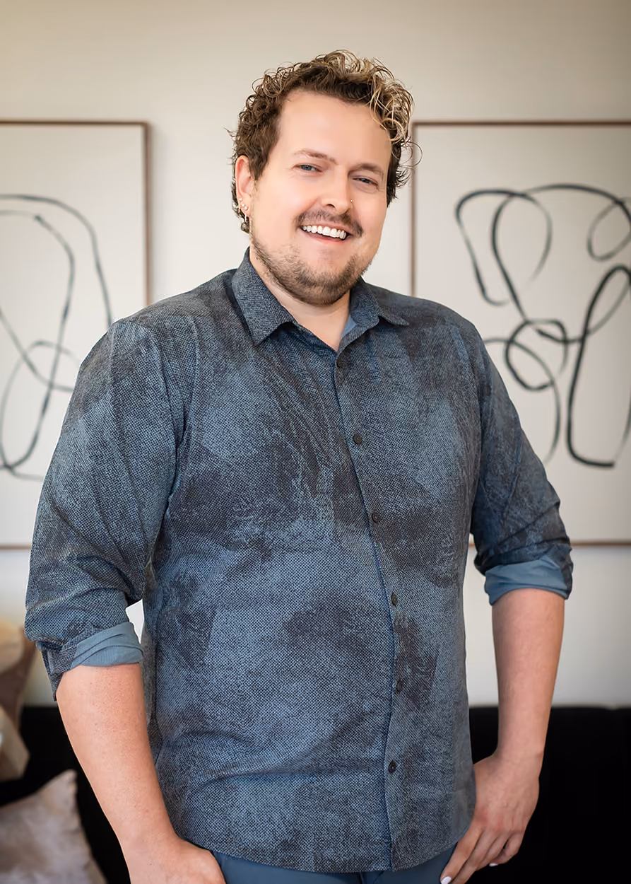 Smiling man with curly hair and a short beard wearing a rolled-up sleeve button-up shirt standing in front of abstract framed wall art in a cozy room.