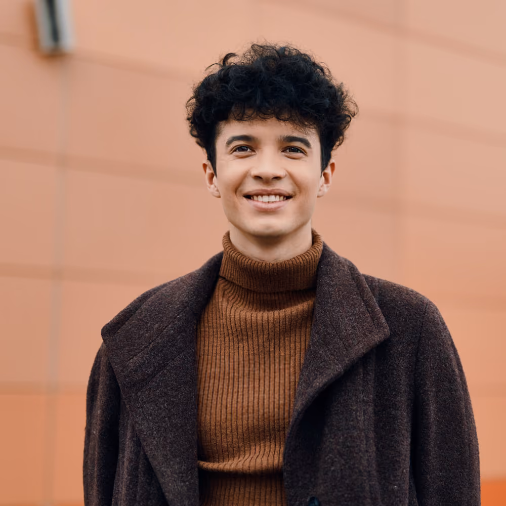 Young man with curly hair smiling, wearing a brown turtleneck sweater and dark brown coat standing in front of a muted peach wall.