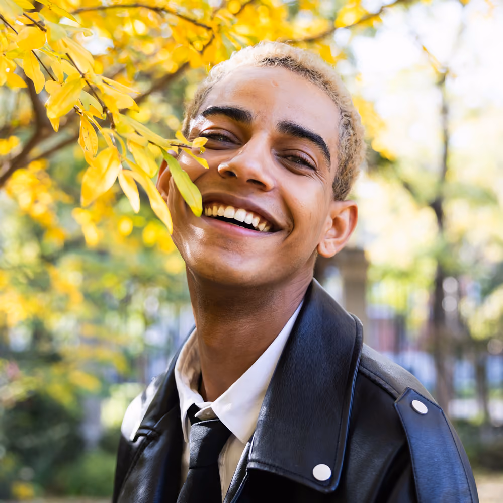 Young man with short blond hair smiling widely, wearing a black leather jacket, white shirt, and black tie, standing outdoors near yellow autumn leaves.
