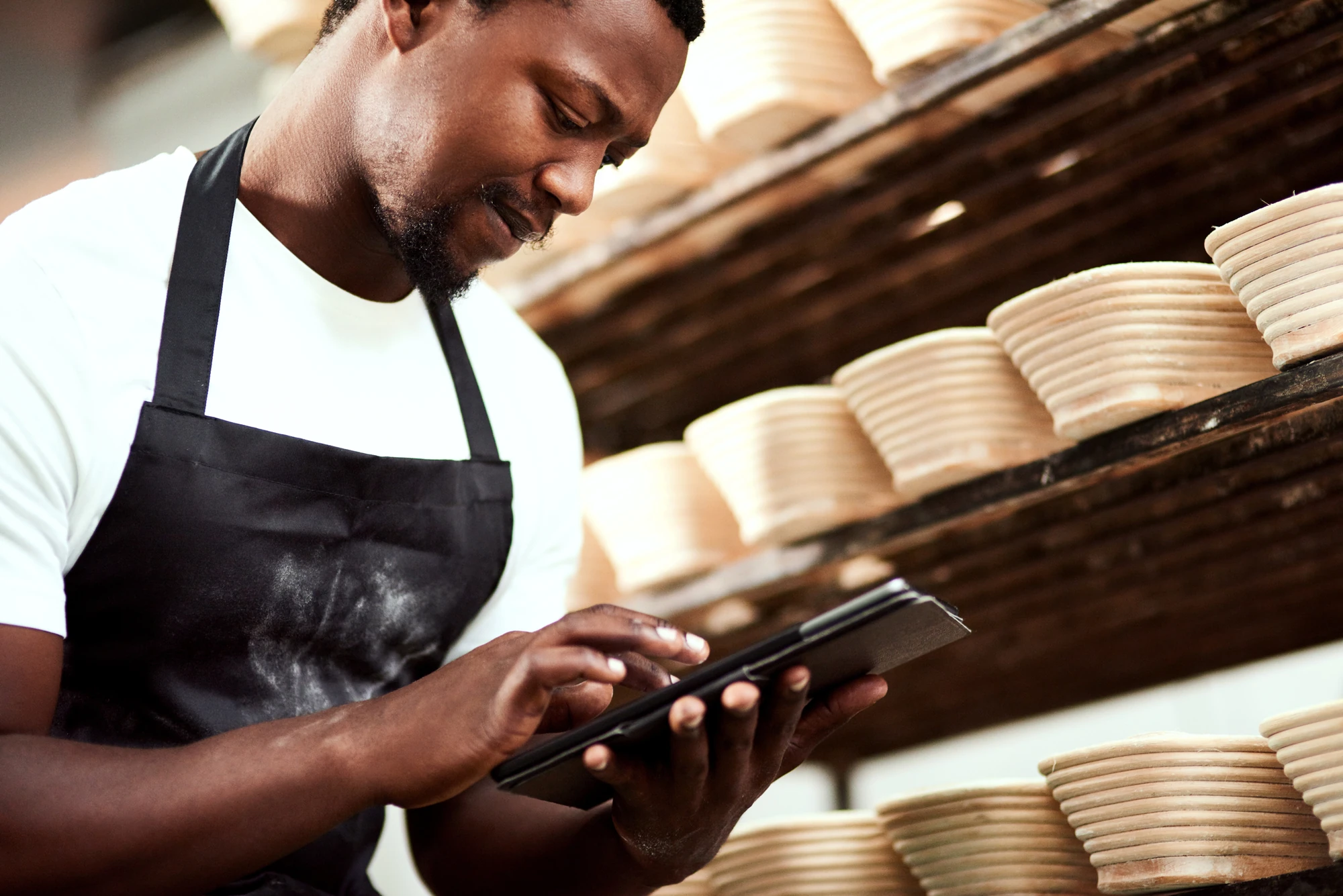 Baker in a flour-dusted apron checking a tablet beside shelves of proofing baskets