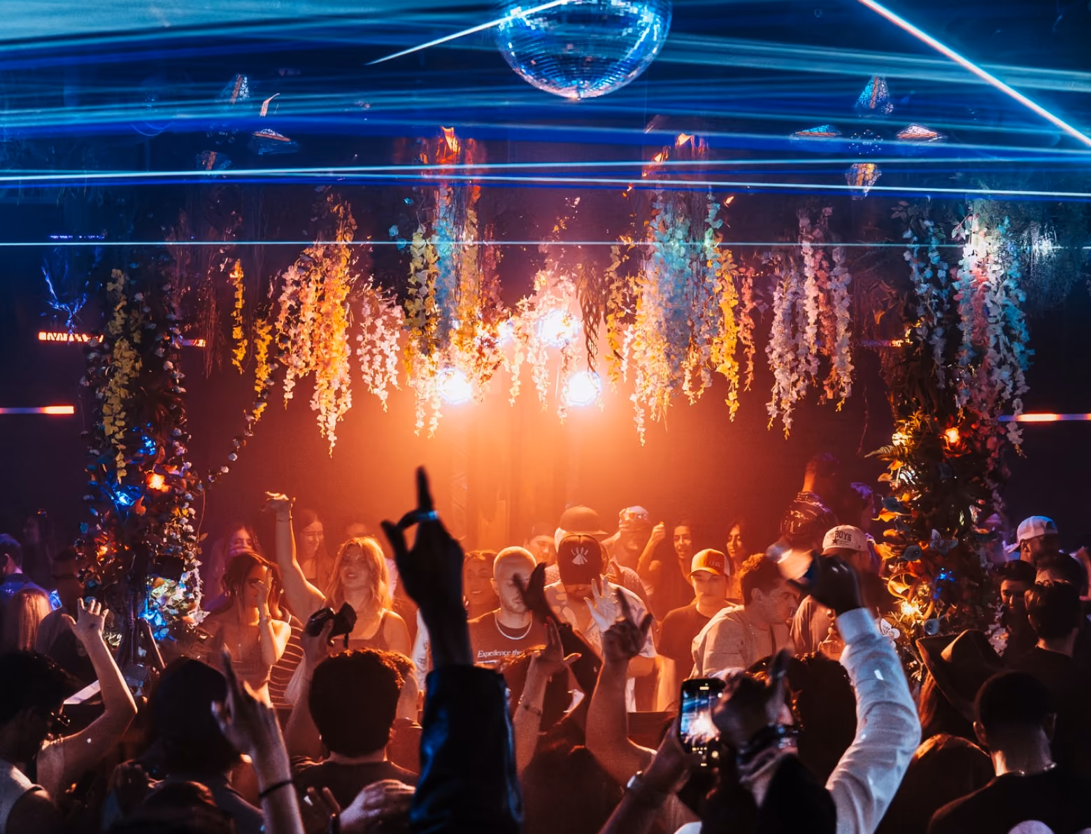 Packed nightclub dance floor under blue laser beams and a disco ball, with hanging floral installations lit orange above the crowd.