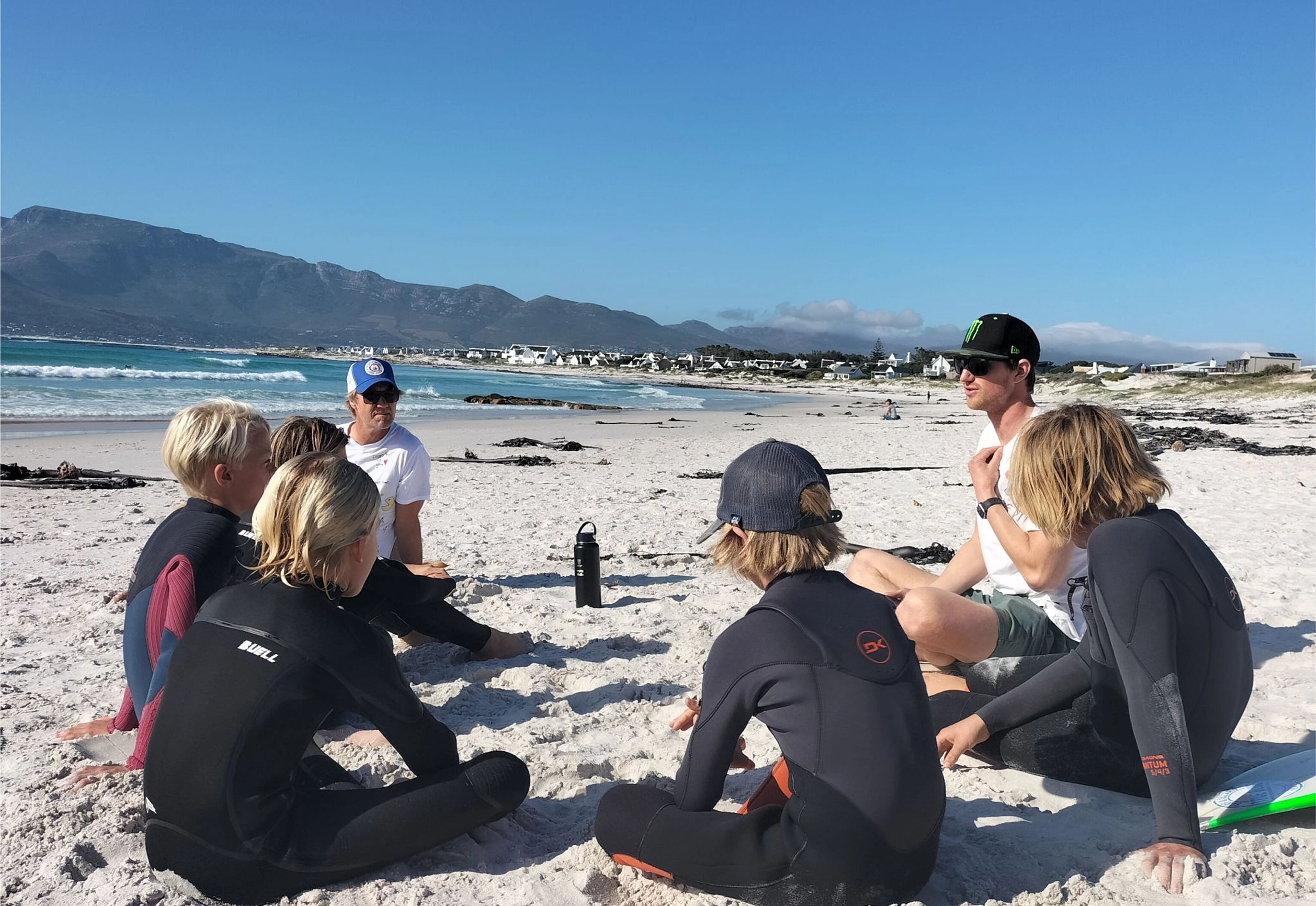 Matt Bromley in a wetsuit sitting on the beach with a group of young surfers