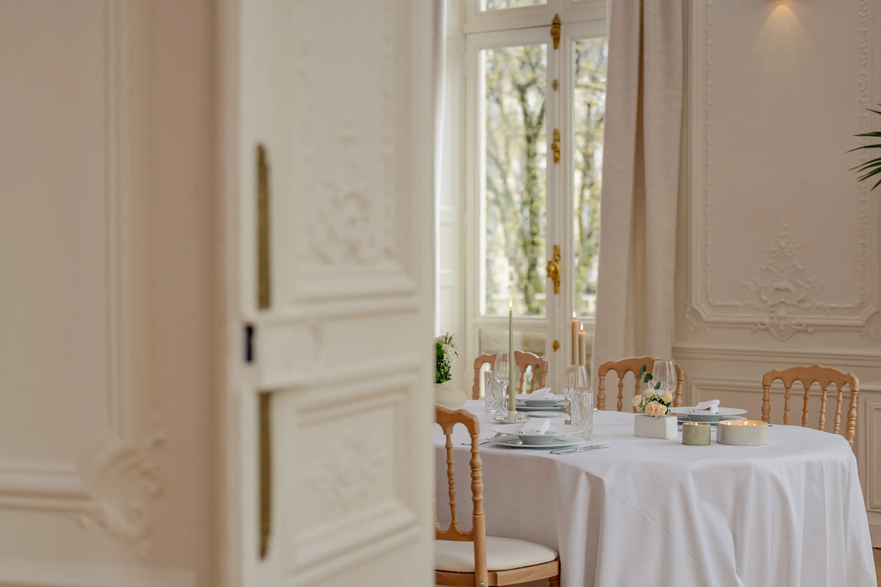Table ronde dressée pour un dîner, chaises Napoléon, bougies et roses blanches devant une fenêtre à la française