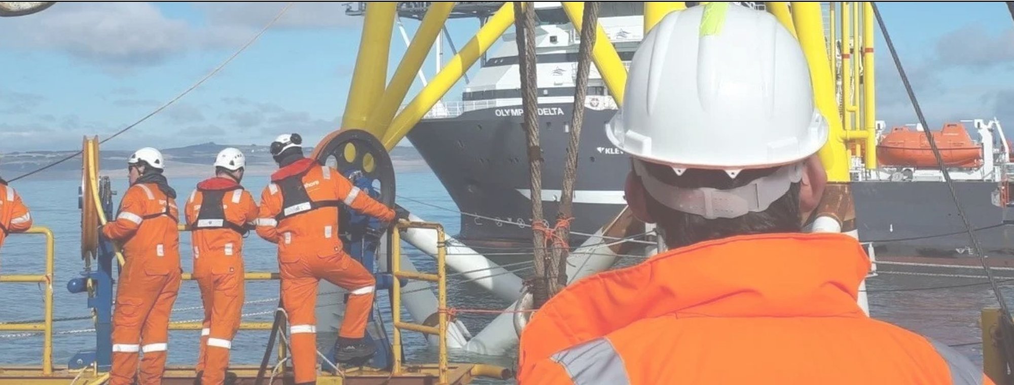 Four workers in orange safety suits and white helmets standing on the deck of an offshore platform near a body of water, with a ship named 'OLYMPIC DELTA' visible in the background.