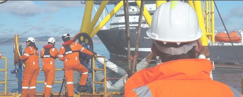 Four workers in orange safety suits and white helmets on an offshore platform, with a large ship visible in the background on a partly cloudy day.