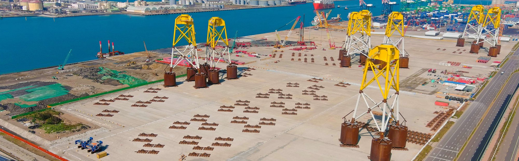 Aerial view of a large industrial port area with several large yellow and white metal structures on concrete ground near a blue body of water, with cranes, containers, and industrial buildings in the background.