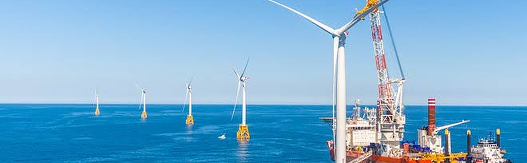 Offshore wind farm with multiple wind turbines in the sea and a large vessel equipped with a crane near the closest turbine under clear blue sky.