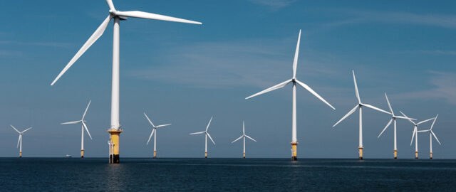 Row of offshore wind turbines standing in the ocean under a clear blue sky.