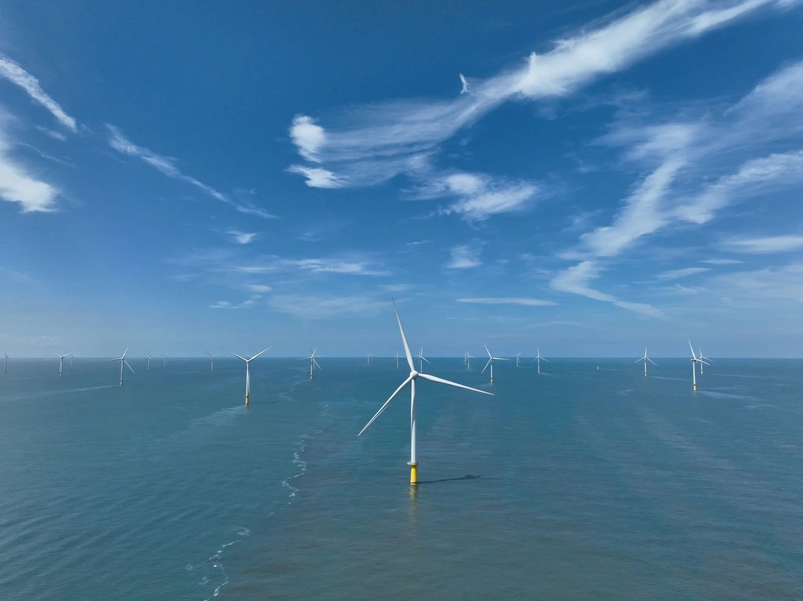 Offshore wind farm with multiple white wind turbines standing in the ocean under a blue sky with wispy clouds.