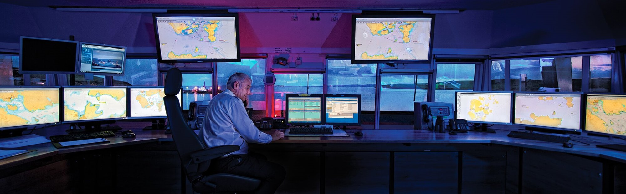 Man sitting in a control room with multiple monitors displaying maps and navigational data, overlooking a waterfront through large windows at dusk.