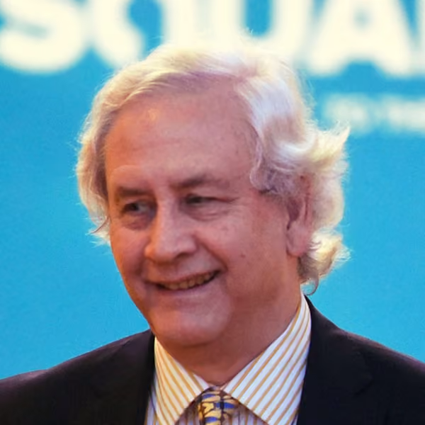 Smiling older man with white hair wearing a striped shirt and patterned tie under a dark jacket, with a blurred blue background.