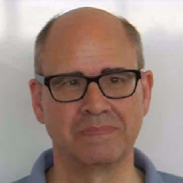 Middle-aged man with a bald head and short hair on the sides, wearing black rectangular glasses and a blue collared shirt, looking slightly to the side with a neutral expression.