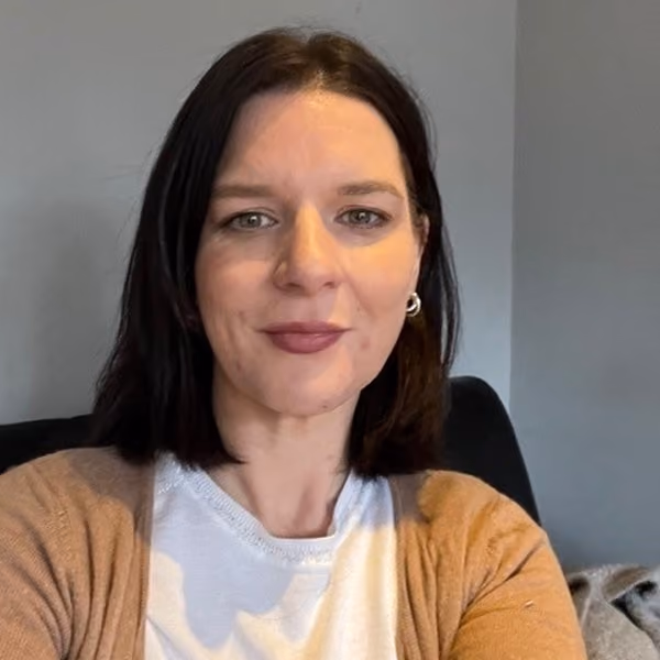 Woman with shoulder-length dark hair, wearing a white shirt and brown cardigan sitting indoors against a plain gray wall.