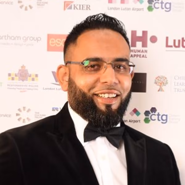 Formal event participant wears black tuxedo with glasses and various sponsor logos displayed.