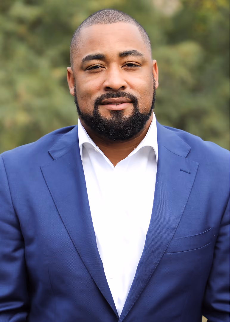 Portrait of a man with closely cropped hair and a beard, wearing a blue suit jacket and white shirt, with a green blurred background.