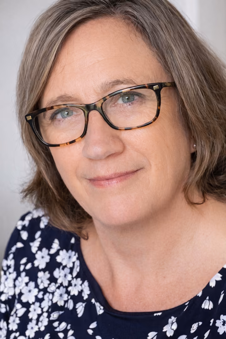 Middle-aged woman with short light brown hair wearing tortoiseshell glasses and a navy blue top with white floral patterns, smiling gently.