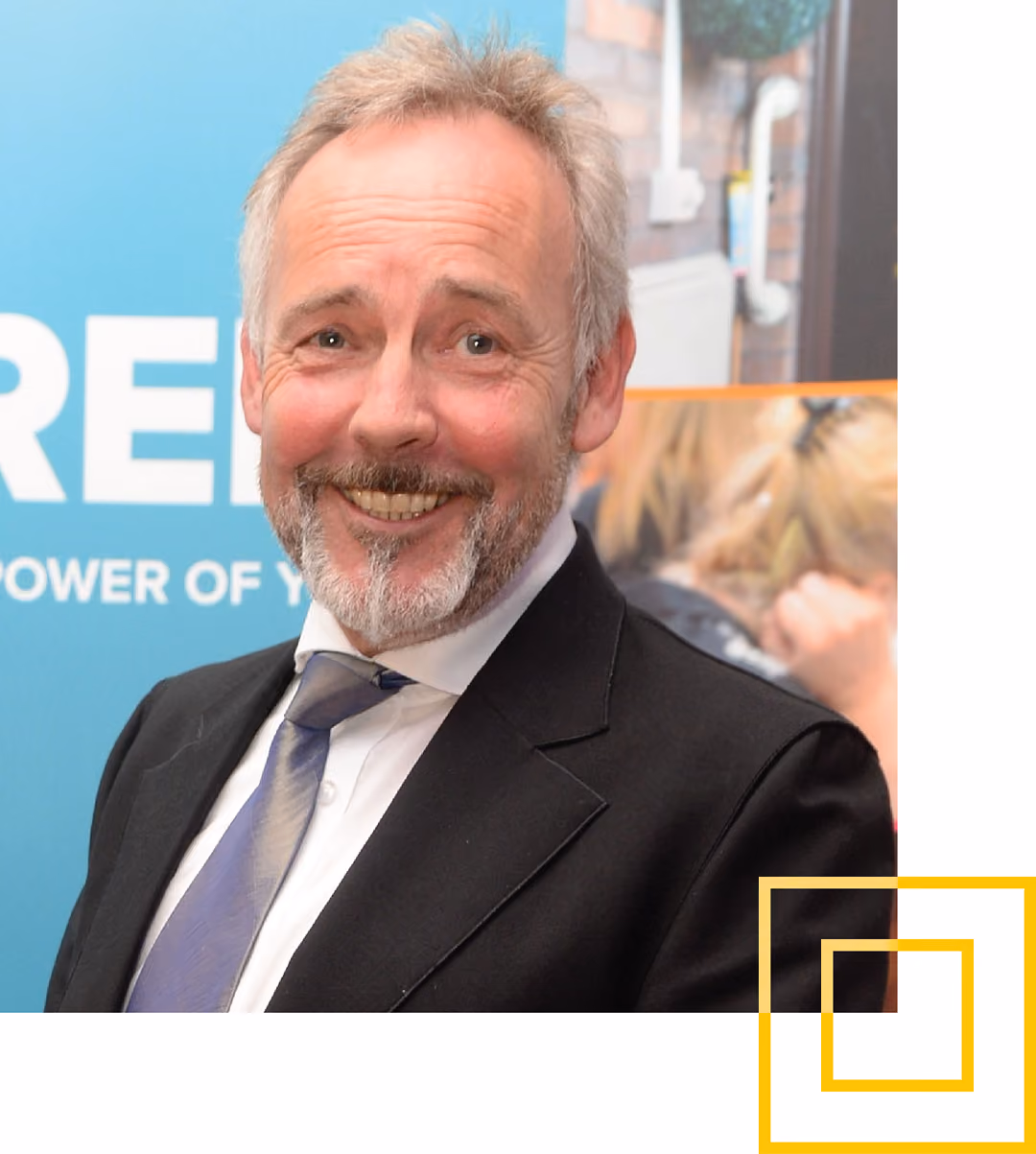 Smiling middle-aged man with gray hair and beard wearing a black suit, white shirt, and blue tie against a blurred background with blue and orange elements.