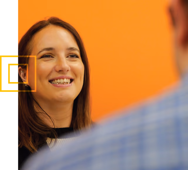 Smiling woman with brown hair and a black top looking at a person in a blue checkered shirt, with an orange background.