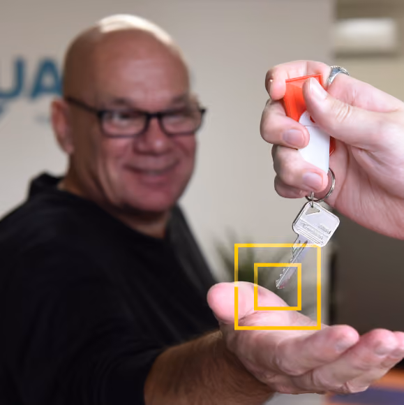 Hand holding a key with a red and white keychain above an open palm of an older man wearing glasses, smiling in the background.