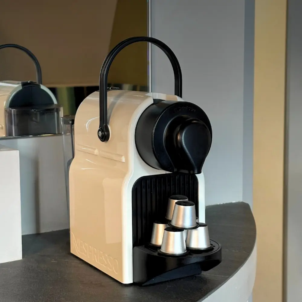 Machine à café Nespresso beige avec poignée noire posée sur un comptoir gris, avec cinq capsules argentées empilées devant l'appareil.