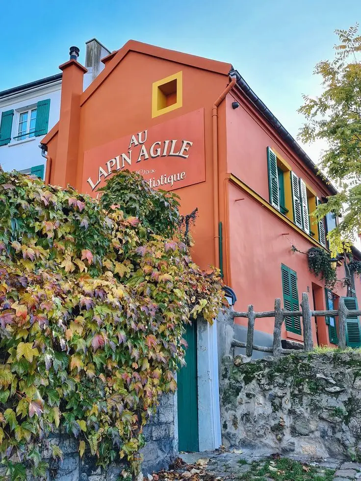 Façade d'un bâtiment coloré en ocre avec des volets verts et une enseigne annonçant 'Au Lapin Agile, Espace Artistique', entouré de végétation et d'un ciel bleu limpide.