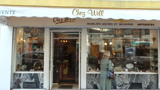 Façade d'une boutique nommée 'Chez Will' avec une enseigne blanche et jaune, affichant 'Mobilier ancien et moderne' et 'Antiquités', une personne âgée en manteau vert observe la vitrine présentant des objets antiques sur fond de murs en marbre noir et blanc.