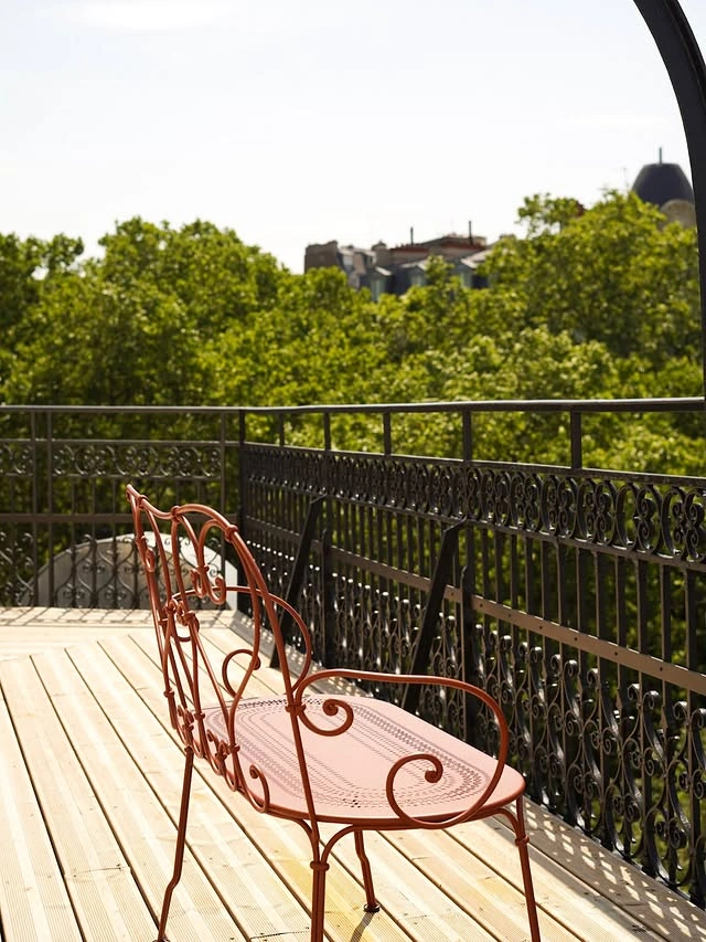 Banc en métal rouge avec motifs décoratifs sur une terrasse en bois, avec une balustrade en fer forgé noire et des arbres verts en arrière-plan.