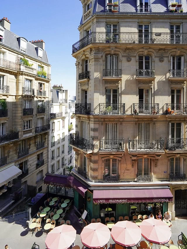 Vue d'un café-restaurant parisien avec des parasols roses, des tables et des chaises alignées en terrasse au pied d'un immeuble haussmannien avec balcons en fer forgé.
