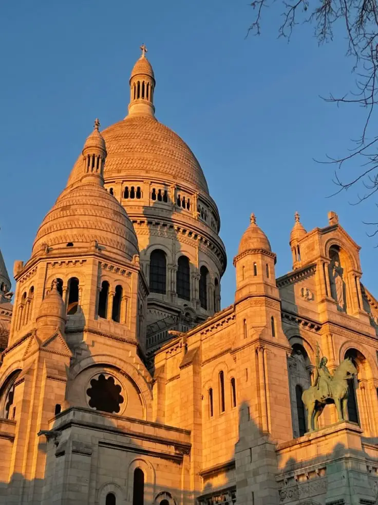 Vue rapprochée des dômes et tours de la basilique du Sacré-Cœur à Paris, éclairés par la lumière dorée du coucher de soleil sur un ciel bleu clair.