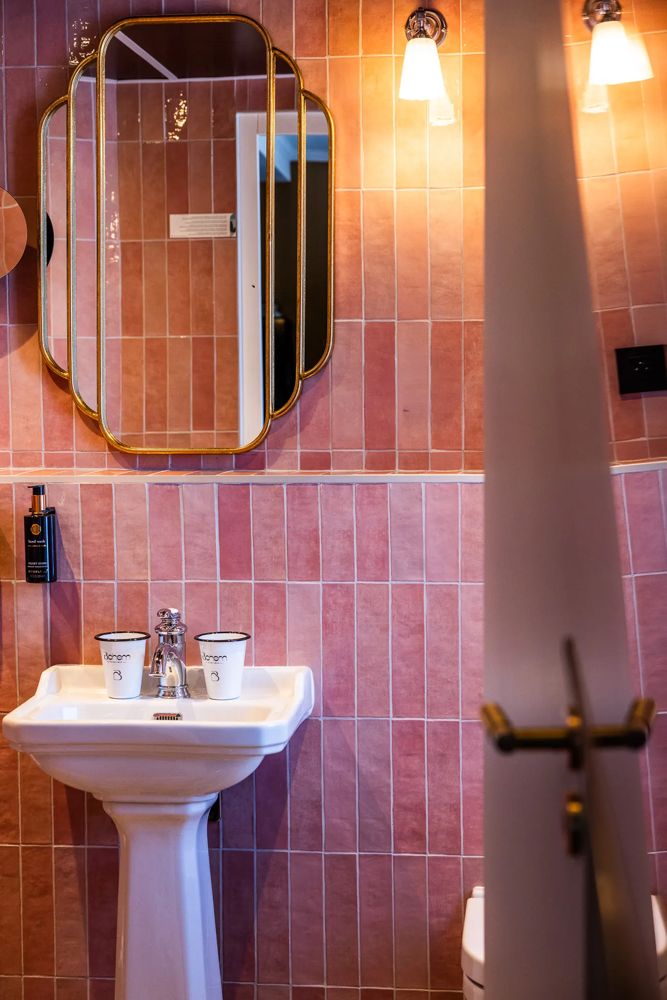 Salle de bain avec un mur carrelé rose, un lavabo blanc sur colonne avec deux gobelets blancs et un miroir décoratif à cadre doré au-dessus, éclairé par une applique murale.