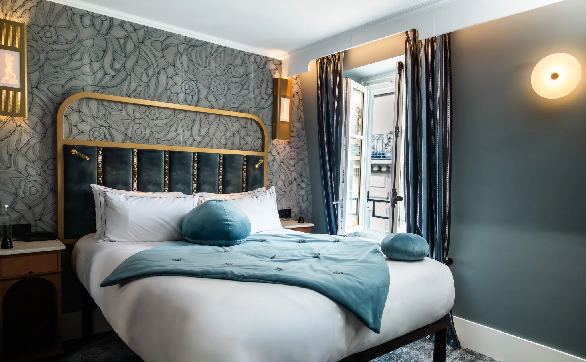 Chambre avec un grand lit blanc, une tête de lit bleue et or, une couverture bleue et des coussins ronds bleus, un mur avec papier peint floral gris, une fenêtre ouverte avec rideaux bleus foncés et une lumière murale ronde allumée.