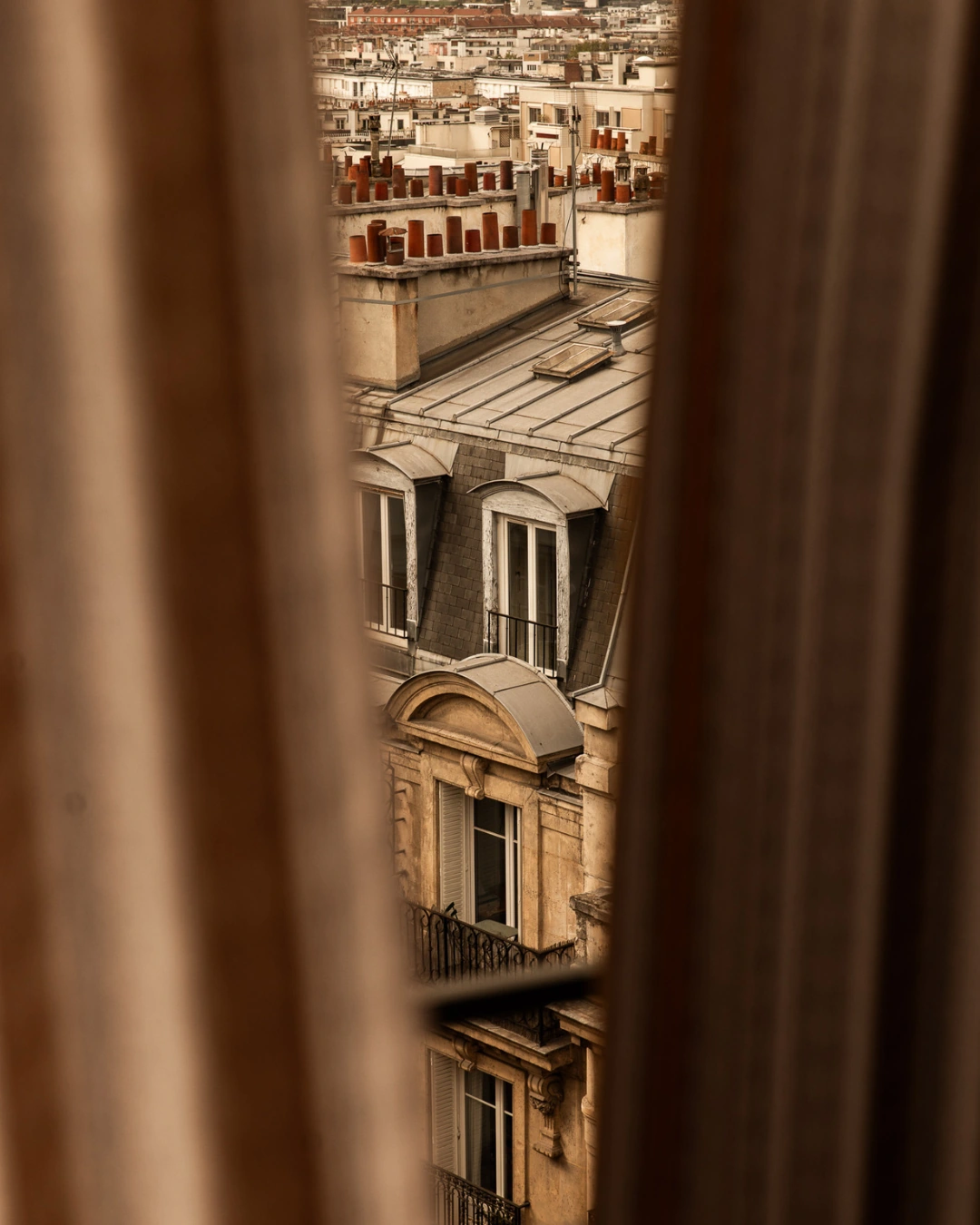 Vue à travers un rideau étroit sur les toits classiques de Paris avec des cheminées en brique, fenêtres mansardées, et les balcons en fer forgé des immeubles anciens.