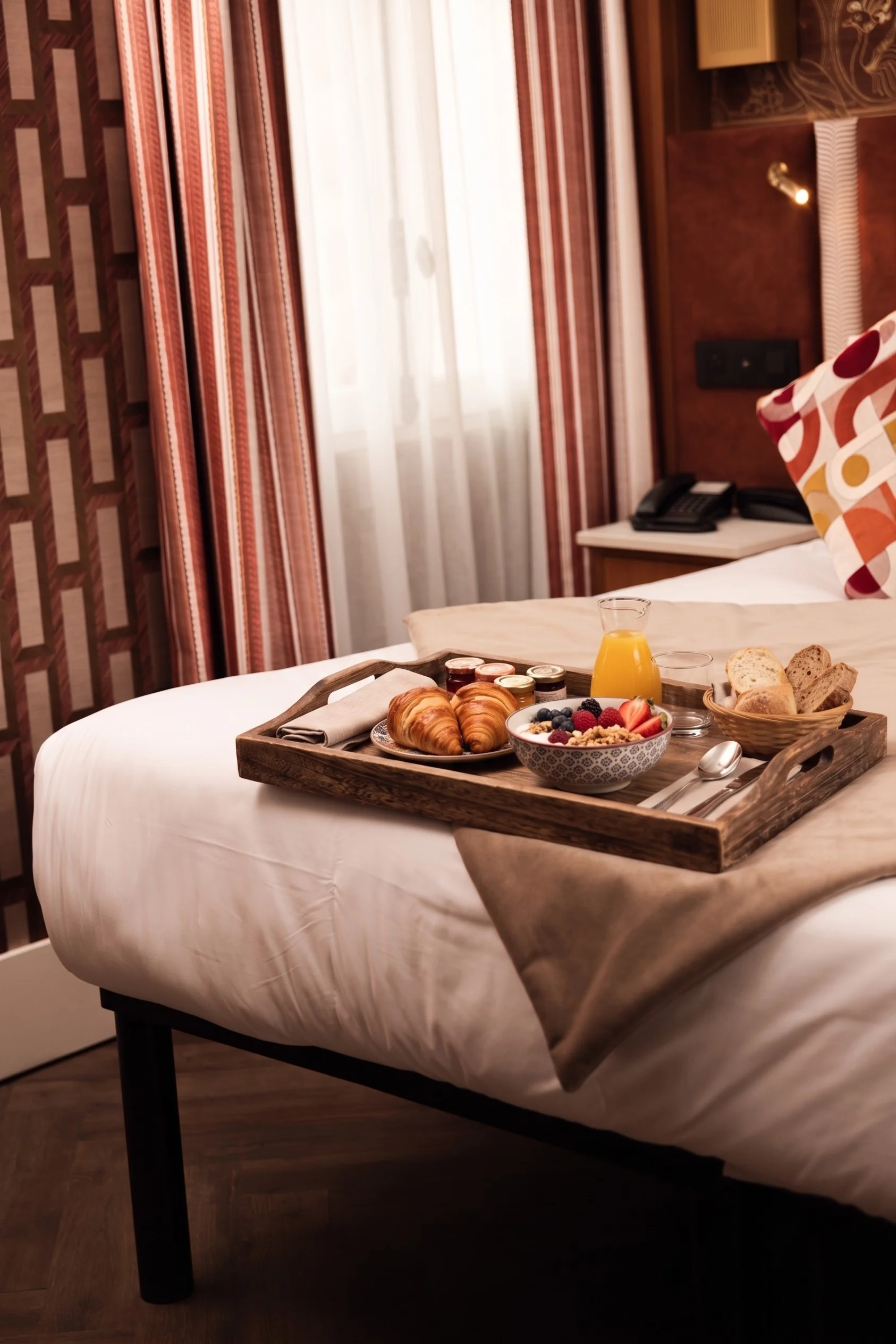 Plateau en bois sur un lit d'hôtel avec deux croissants, un bol de fruits rouges et granola, un pichet de jus d'orange, un verre, un panier de tranches de pain, quatre petits pots de confiture, des couverts et une serviette.