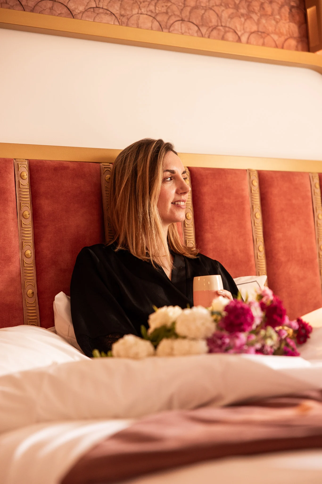 Femme souriante assise dans un lit à tête de lit rouge avec des bandes décoratives dorées, tenant une tasse, avec des fleurs blanches et roses floues au premier plan.