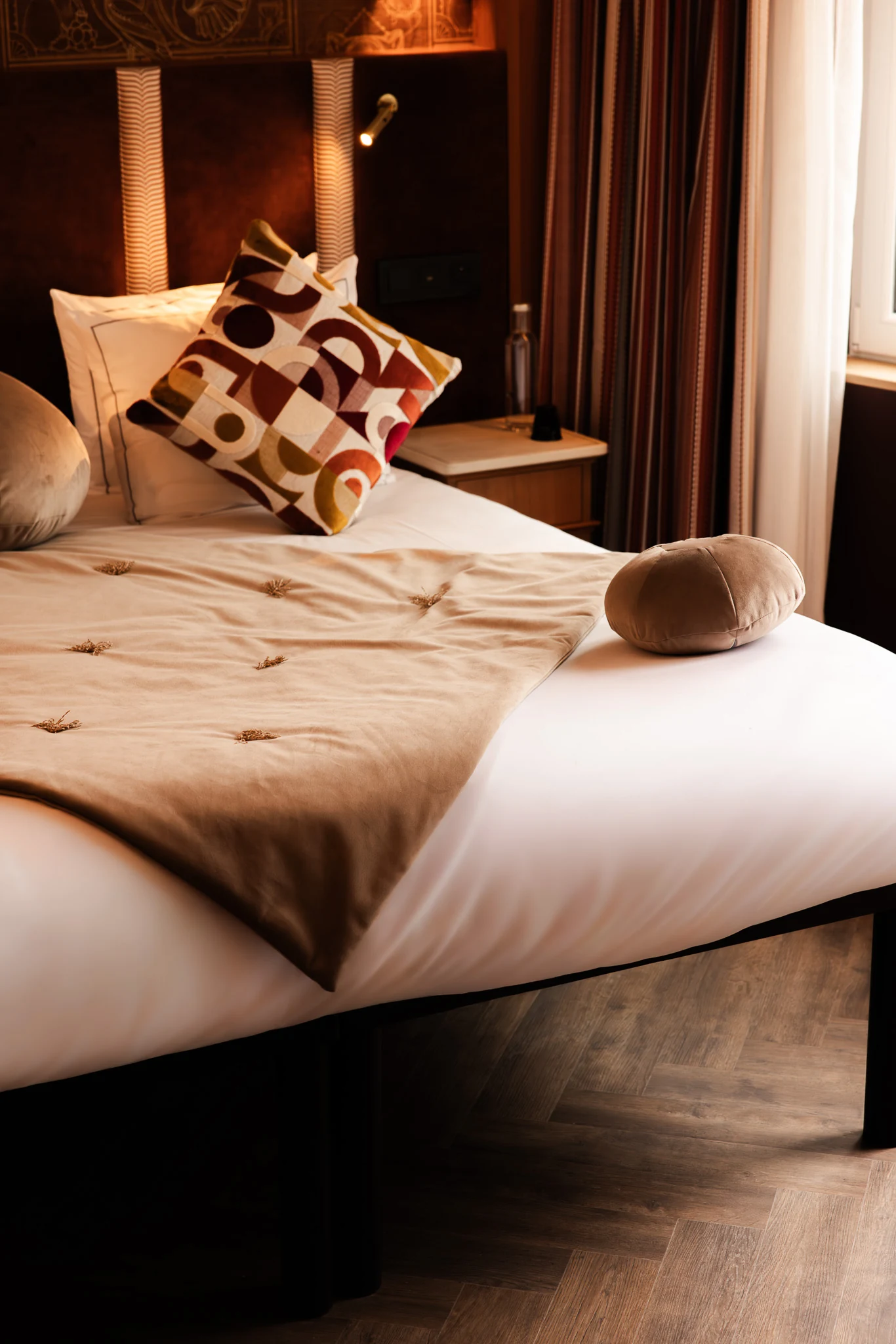 Lit décoré avec une housse beige brodée, un coussin décoratif à motifs géométriques dans des tons marron et un coussin rond assorti, près d'une table de nuit en bois avec une lampe et une bouteille d'eau, à côté d'une fenêtre avec rideaux.