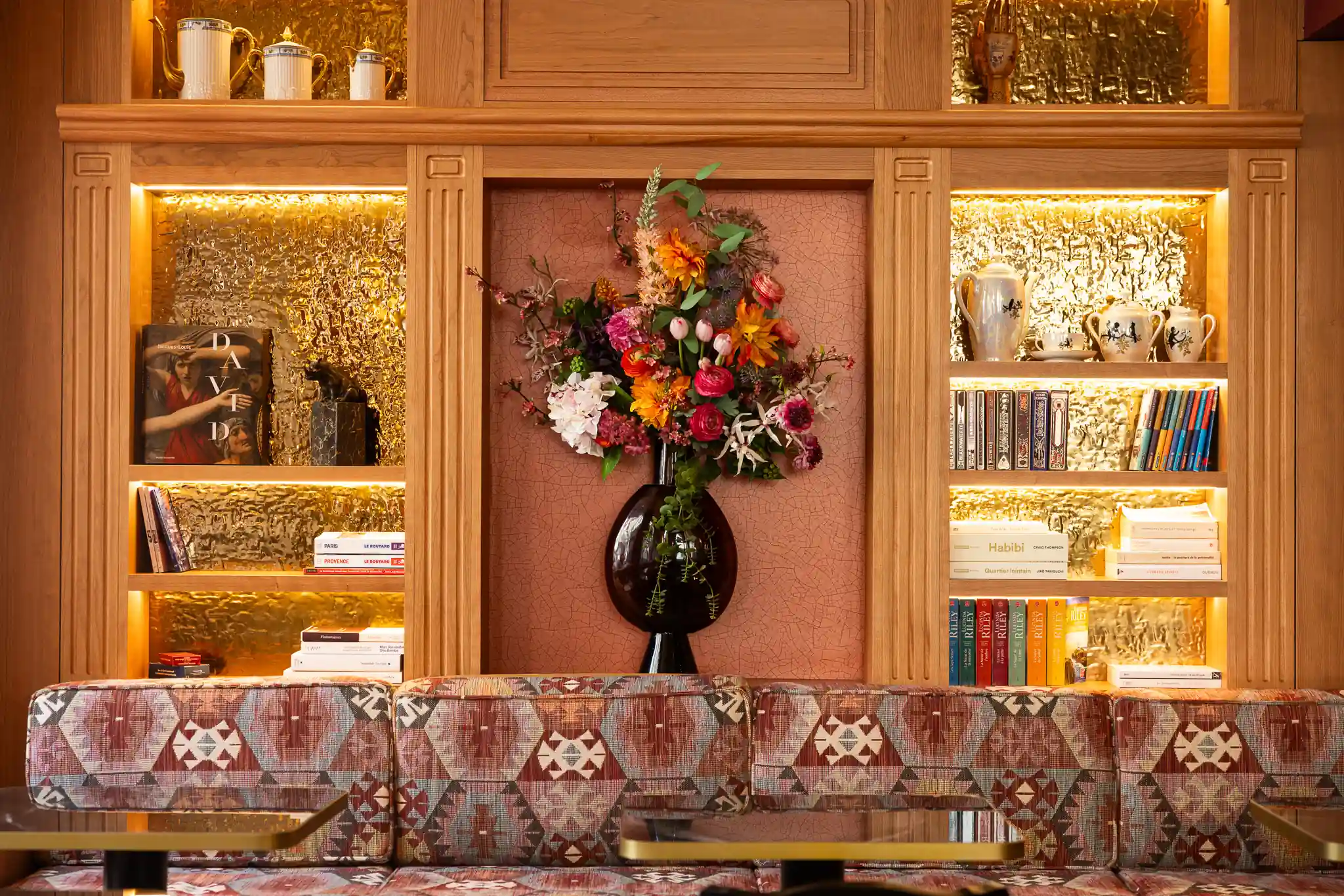 Étagères en bois éclairées avec derrière un mur doré texturé, une grande vase noire au centre contenant un bouquet vif de fleurs multicolores, entourée de livres et d'objets en porcelaine blanche, devant un canapé recouvert de tissu à motifs géométriques rouges et beige, avec deux tables basses en verre et métal.