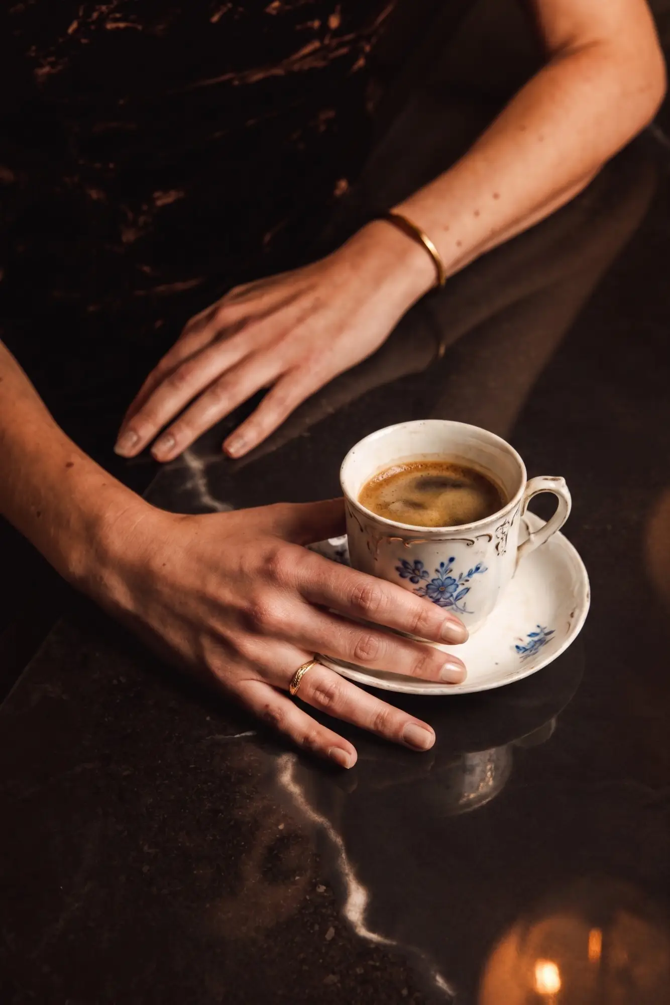 Deux mains féminines posées sur une surface sombre, l'une tenant une tasse en porcelaine blanche décorée de fleurs bleues contenant un café.