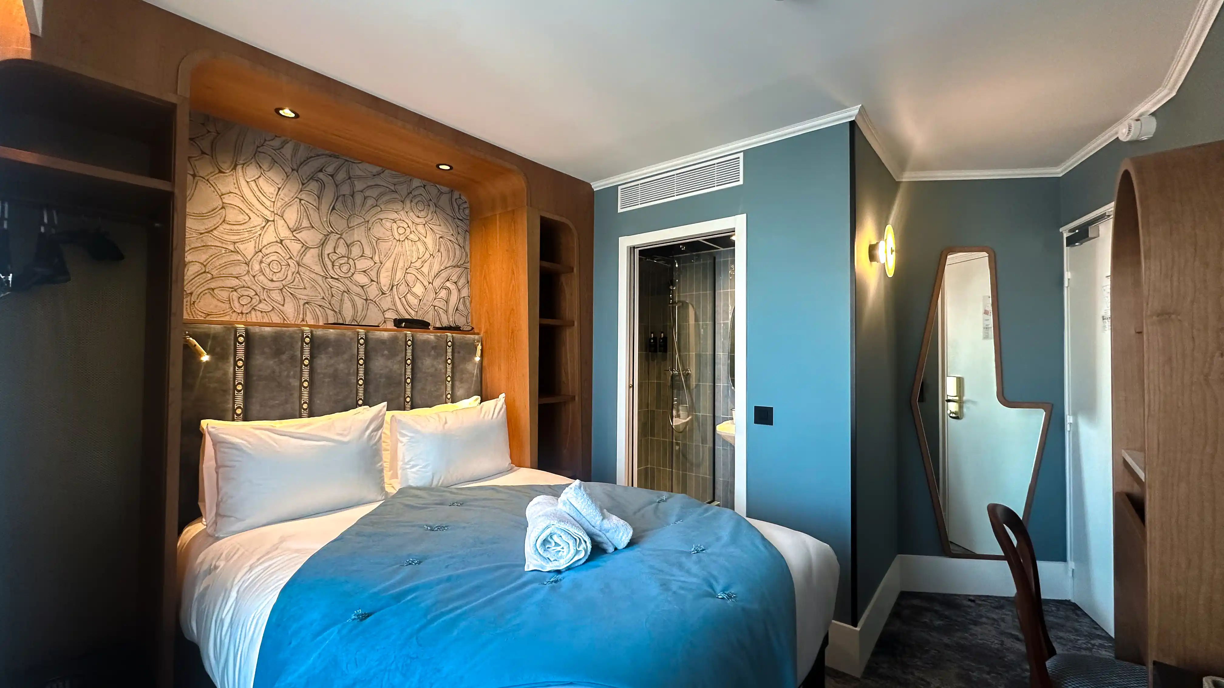 Chambre d'hôtel cosy avec un lit double fait, parure de lit bleu, deux serviettes roulées au centre, tête de lit rembourrée décorée, mur bleu clair avec une porte de salle de bain à carreaux gris, miroir original encadré en bois, chaise et meuble en bois clair.
