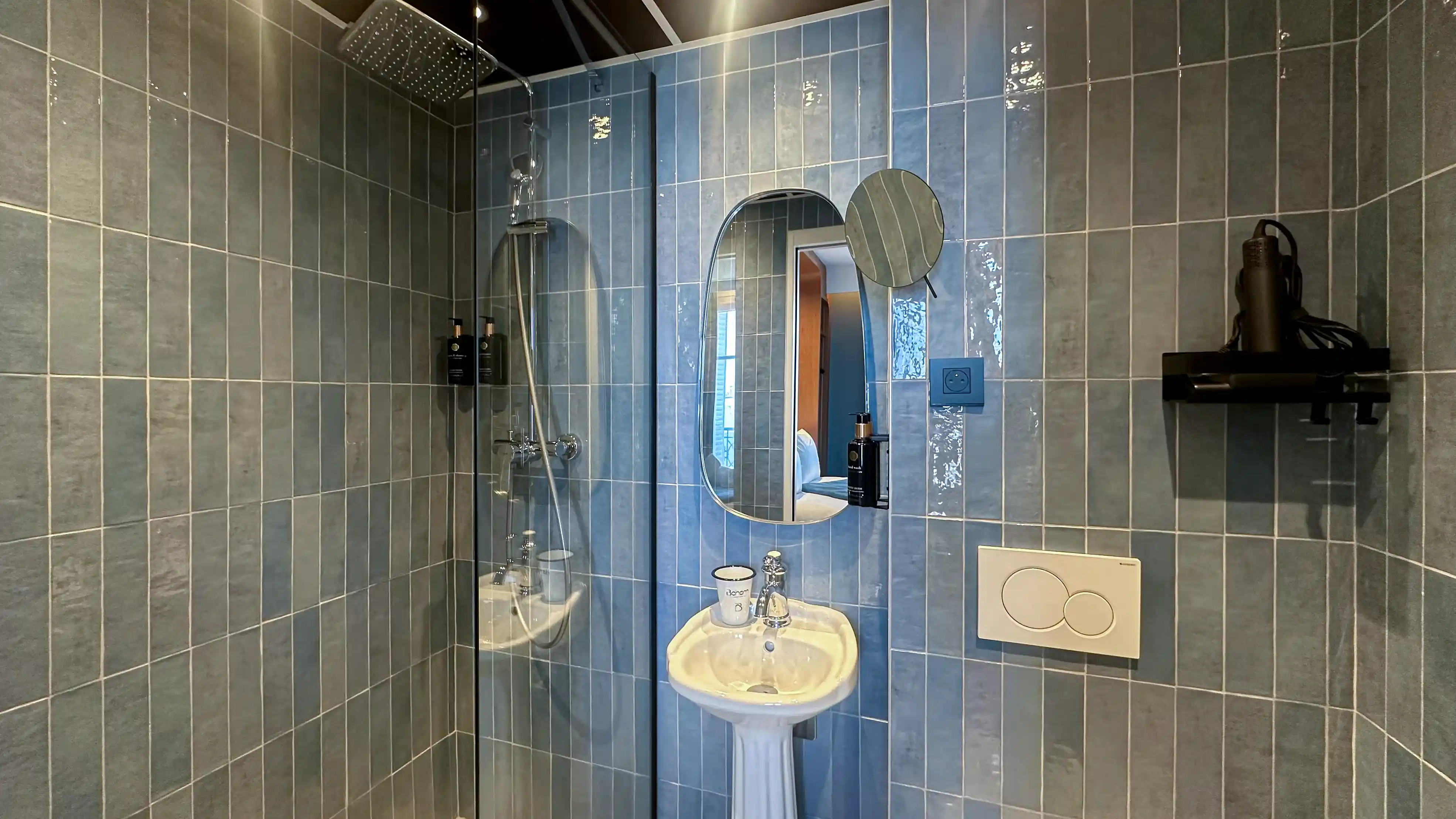 Salle de bain moderne avec murs carrelés bleus, grande douche à tête carrée, lavabo blanc et miroir ovale, sèche-cheveux sur une étagère.