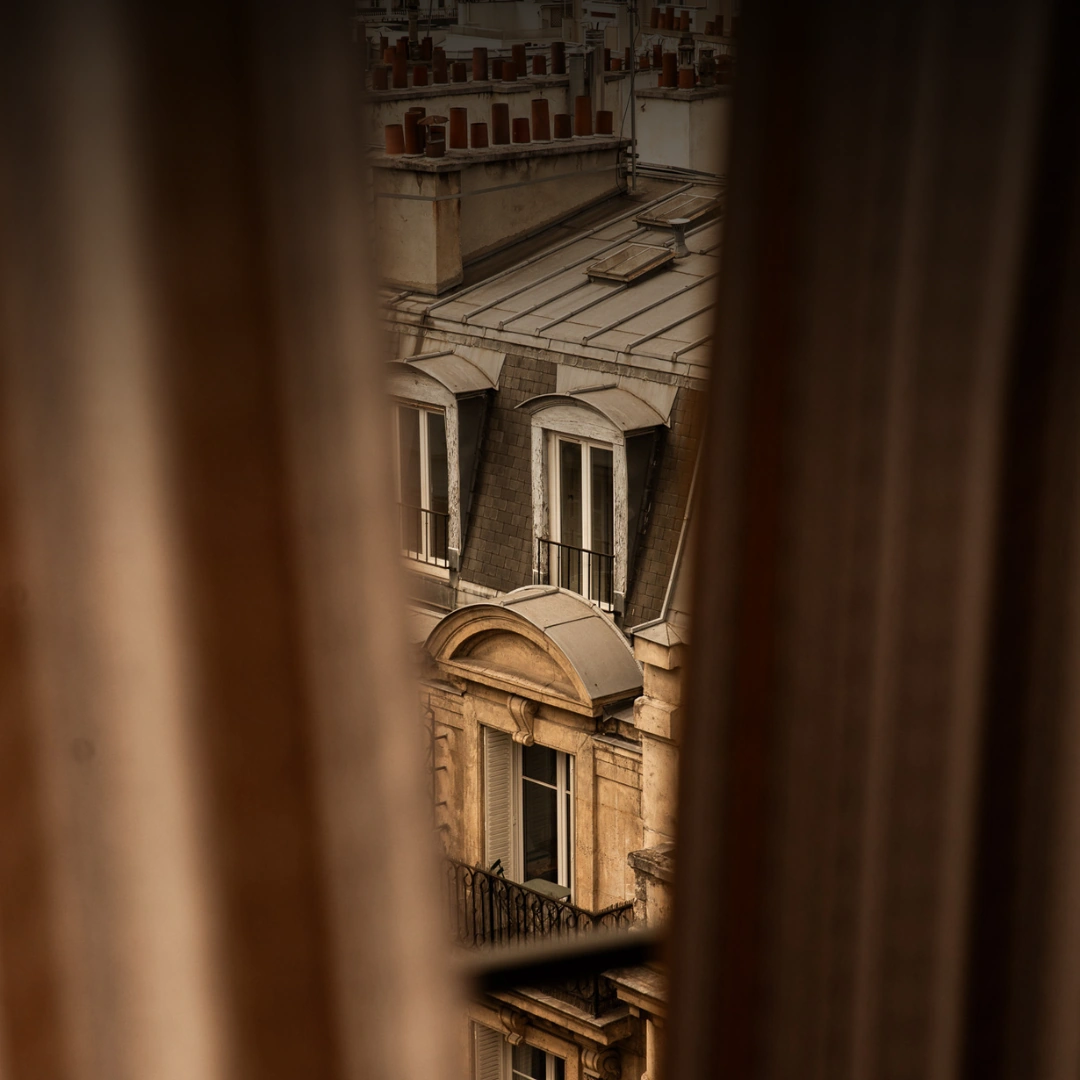 Vue partielle d'un bâtiment parisien ancien avec fenêtres mansardées et toits en zinc, prise à travers un rideau brun flou.