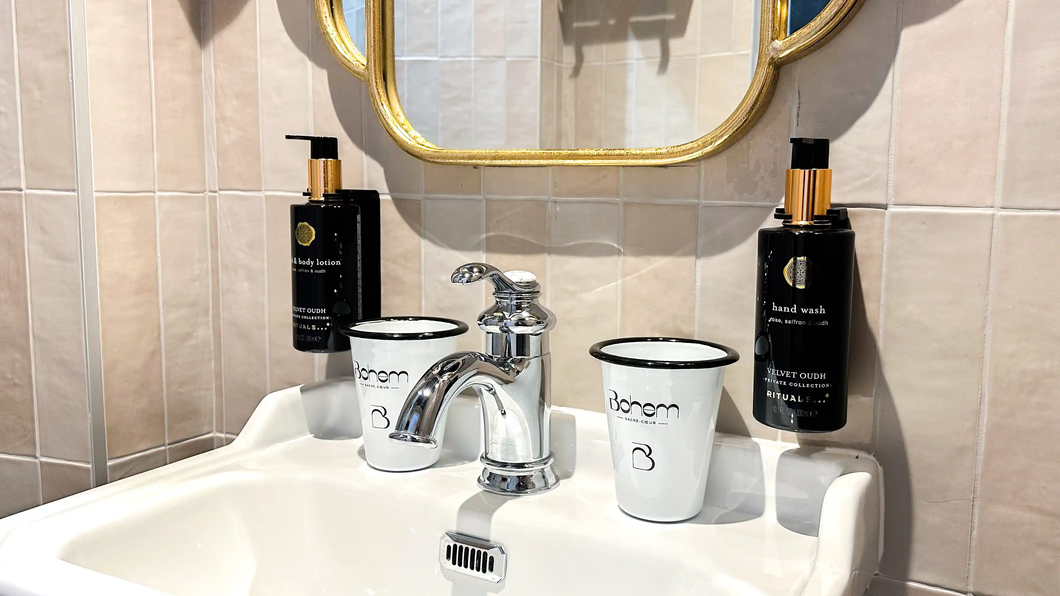 Lavabo blanc avec robinet en métal chromé, deux gobelets blancs avec inscription Bohem et deux distributeurs noirs de lotion pour les mains et le corps de la collection Velvet Oudh de Rituals fixés au mur avec miroir doré en haut et carrelage beige en arrière-plan.