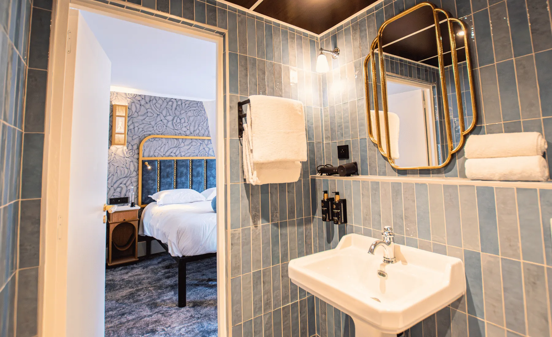 Salle de bain avec murs recouverts de carreaux bleus, lavabo blanc, miroir doré, serviettes blanches pliées et sèche-cheveux. La porte ouverte montre une chambre avec un lit à tête de lit bleue encadrée de doré et un mur avec motif floral.
