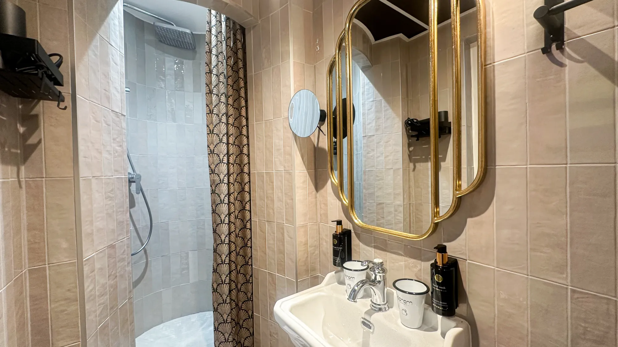 Salle de bain classique avec carrelage beige, lavabo blanc avec miroir doré, deux gobelets, distributeur de savon noir, miroir grossissant et douche avec rideau à motifs noirs et dorés.