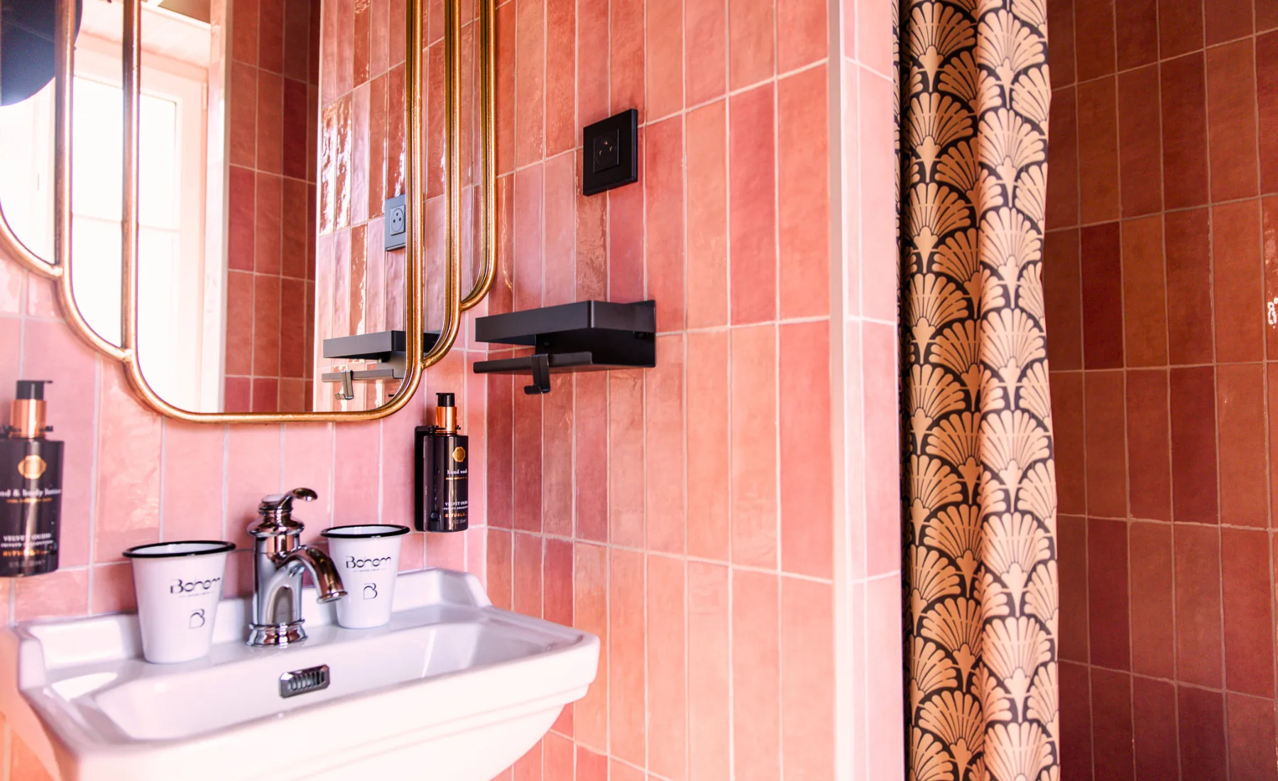 Coin lavabo de salle de bain avec carreaux roses, un miroir à cadre doré, un robinet chromé, deux gobelets blancs marqués Bohem et un rideau de douche art déco à motifs coquilles beige et noirs.