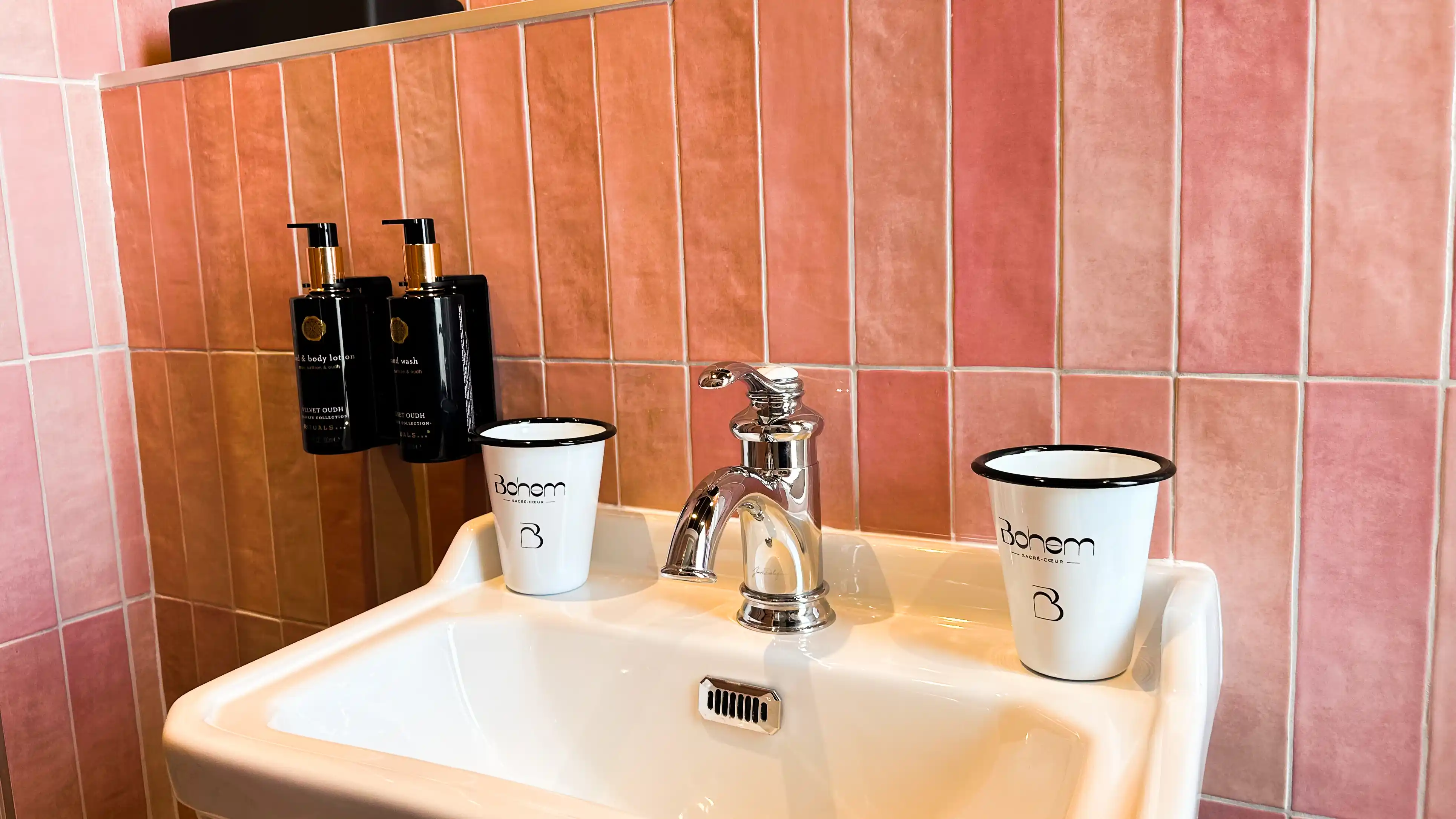 Évier blanc avec robinet en métal brillant, deux gobelets blancs avec logo Bohem et deux distributeurs noirs de savon montés sur un carrelage mural de couleur rose.