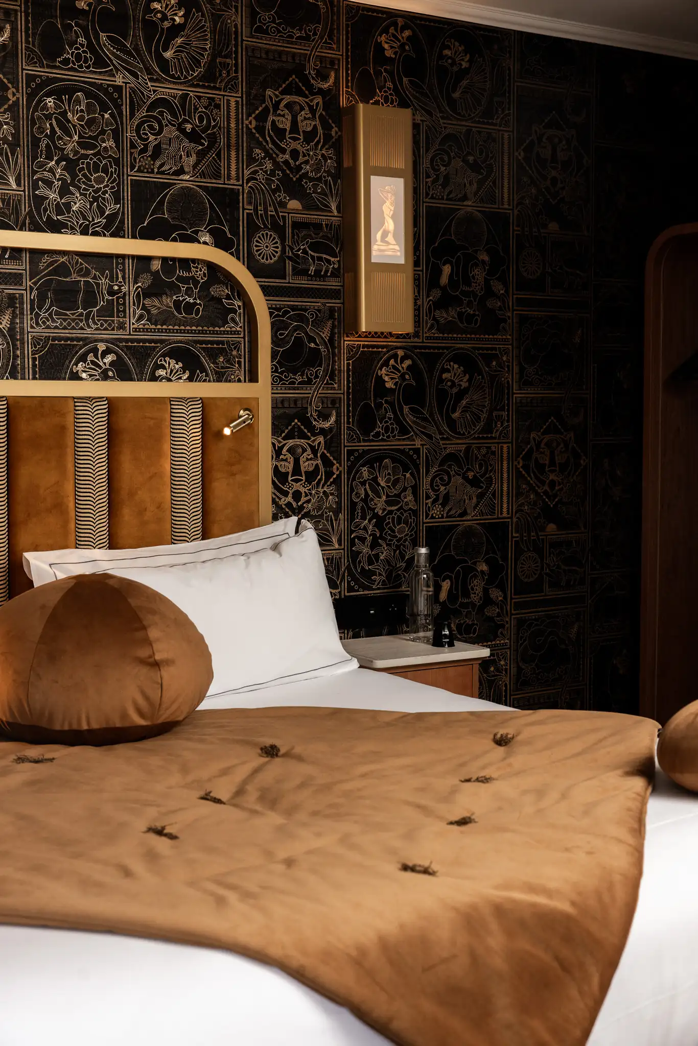 Chambre avec un lit habillé d'une couverture et coussins ronds en velours camel, tête de lit assortie, et mur décoré de motifs animaux dorés sur fond noir.