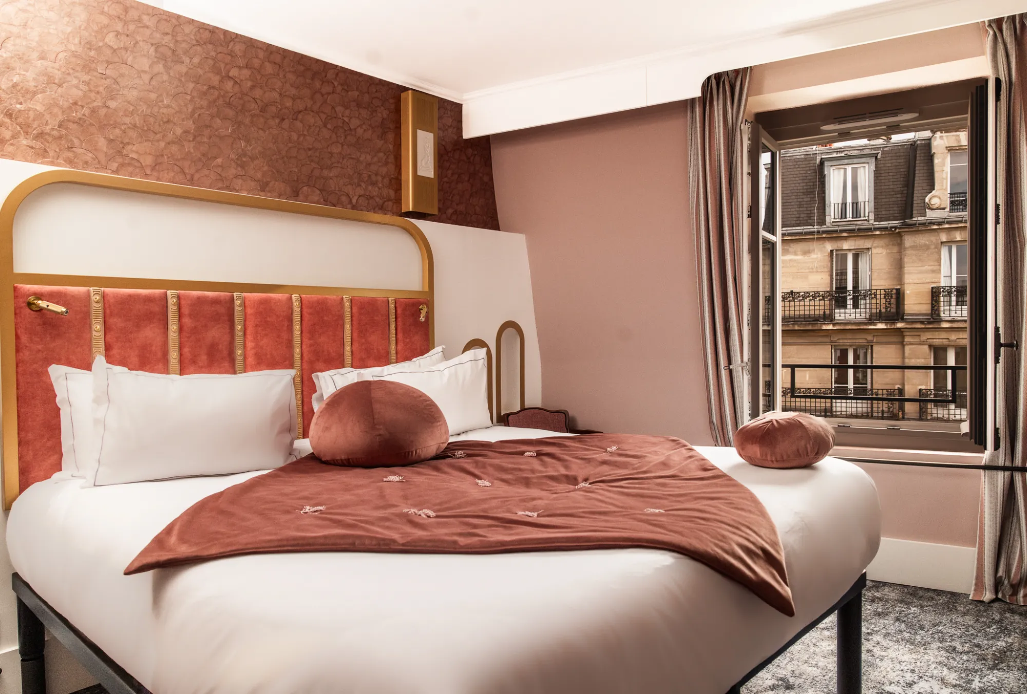 Chambre avec lit double aux draps blancs, tête de lit rouge avec cadre doré, couverture et coussins ronds couleur terracotta, fenêtre ouverte donnant sur un immeuble en pierre avec balcons en fer forgé.