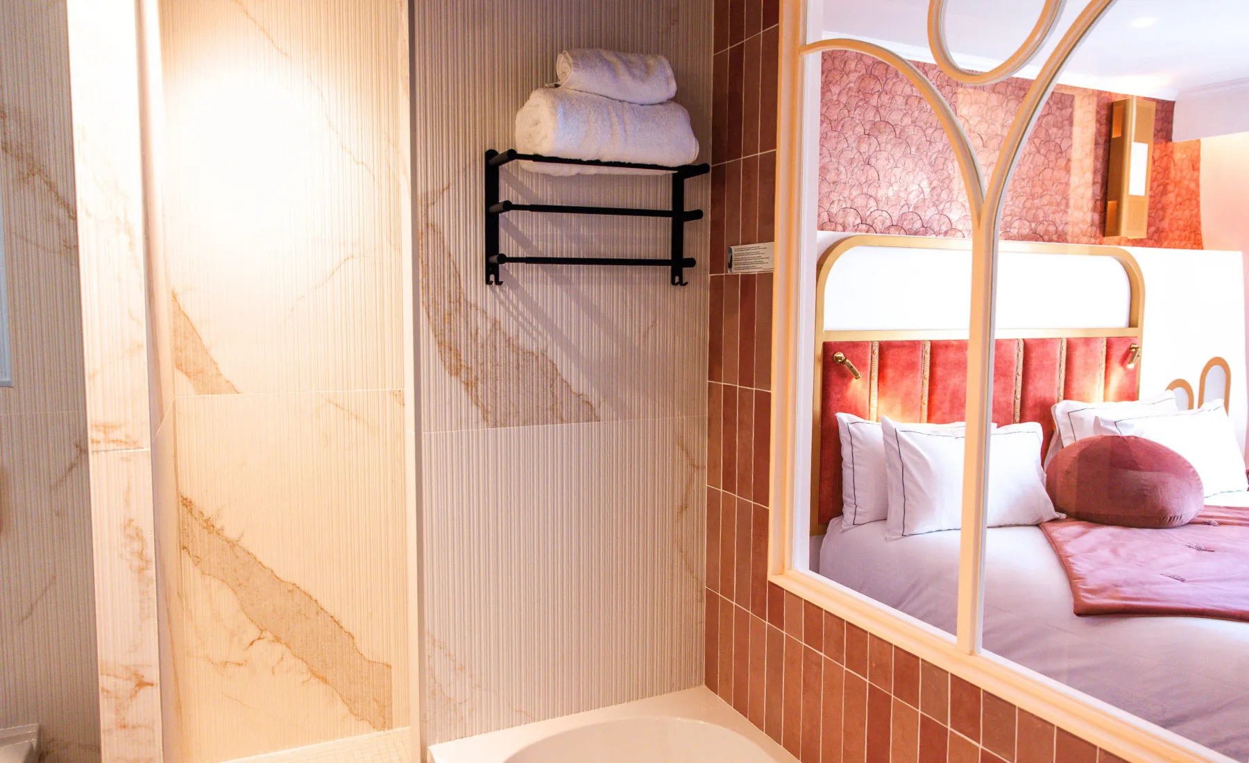 Salle de bain avec murs carrelés beige marbré et porte-serviettes noir avec serviettes blanches, avec une fenêtre donnant sur une chambre à coucher décorée de murs roses, un lit avec une tête de lit rembourrée rose, des oreillers blancs et un plaid rose.