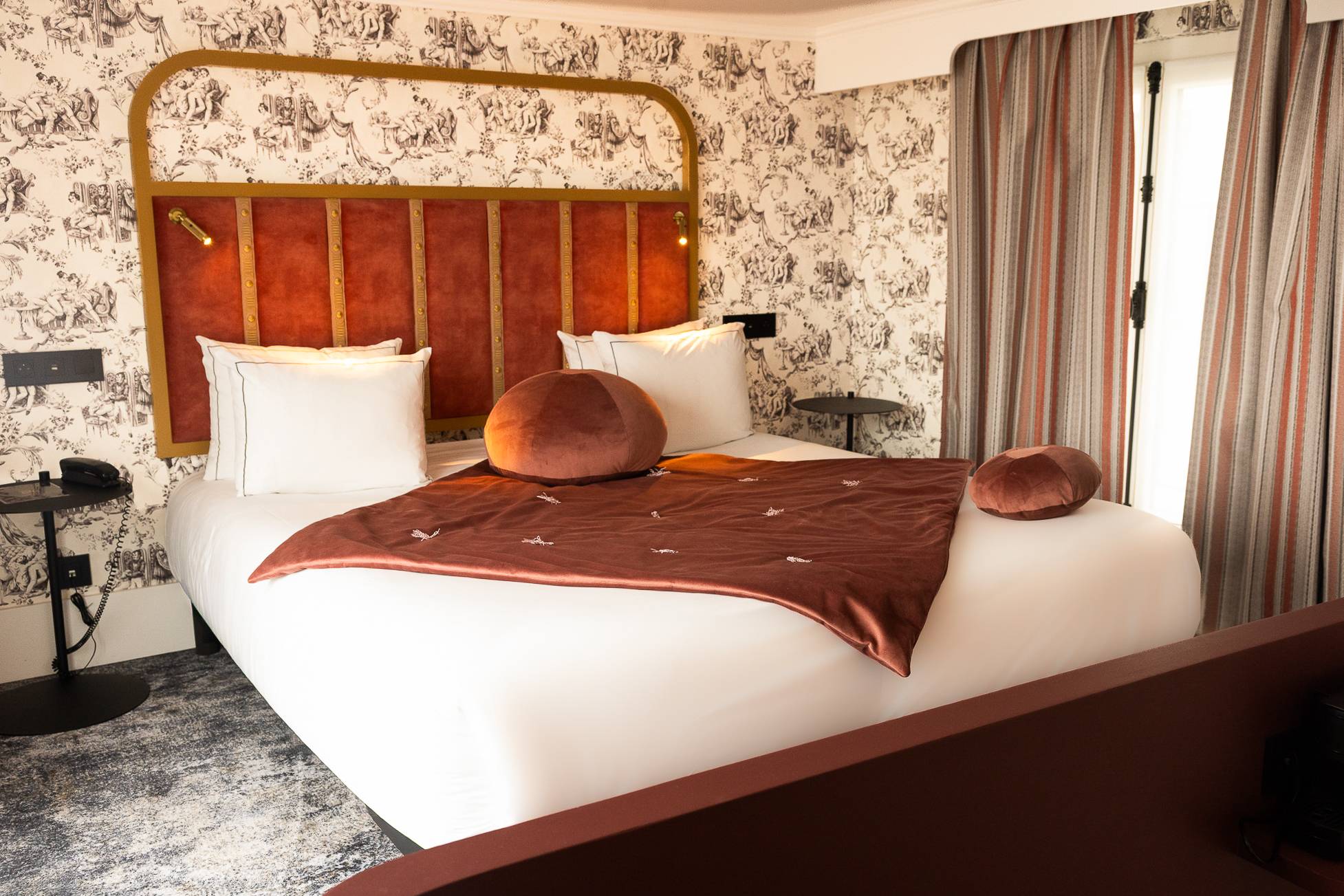 Chambre avec un grand lit double avec une tête de lit en velours rouge encadrée de métal doré, deux oreillers blancs, deux coussins ronds en velours rouge, une couverture rouge avec petits motifs blancs, des tables de chevet noires, un téléphone, des rideaux à rayures rouges et beiges, et un mur décoré de papier peint noir et blanc à motifs classiques.